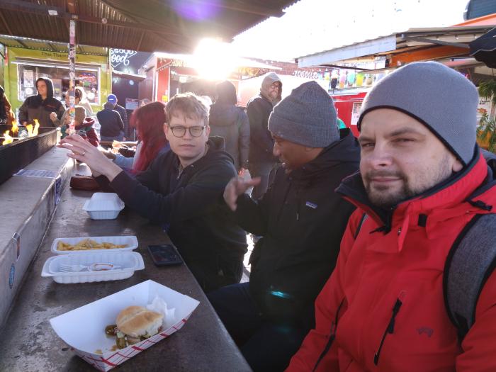 Three people eating in a busy foot court outside.