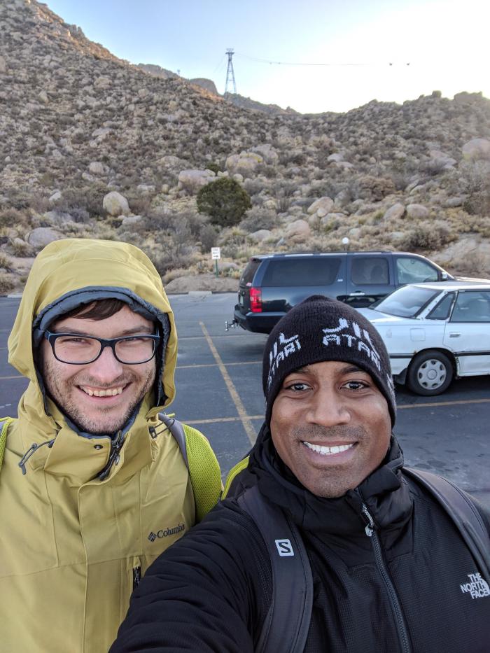A selfie of two male individuals with two parked cars and a desert in the background.
